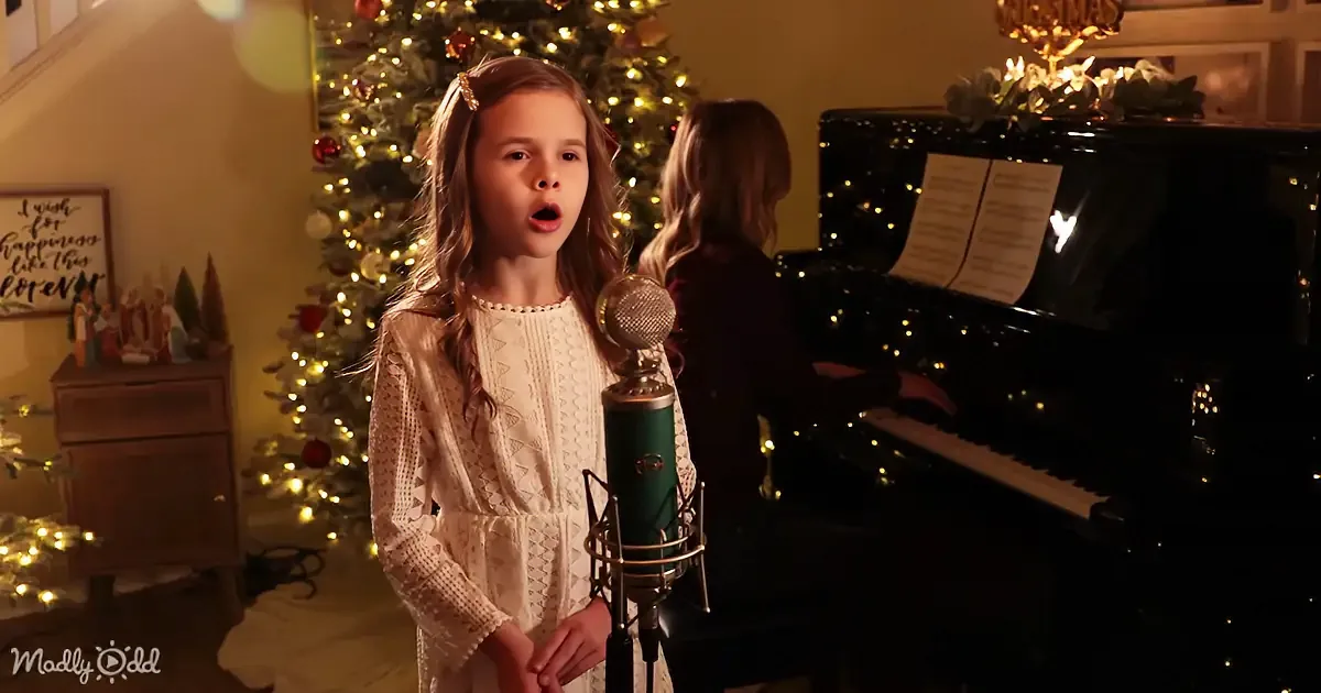A young girl sings into a microphone in front of a Christmas tree and piano.