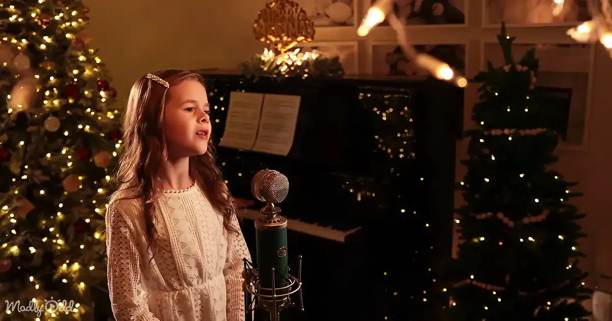 A young girl in a white dress sings into a microphone, surrounded by Christmas trees and lights.