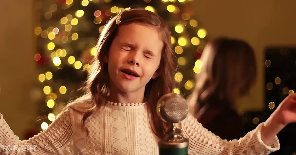 A young girl in a white sweater sings into a microphone, with a Christmas tree and lights in the background.