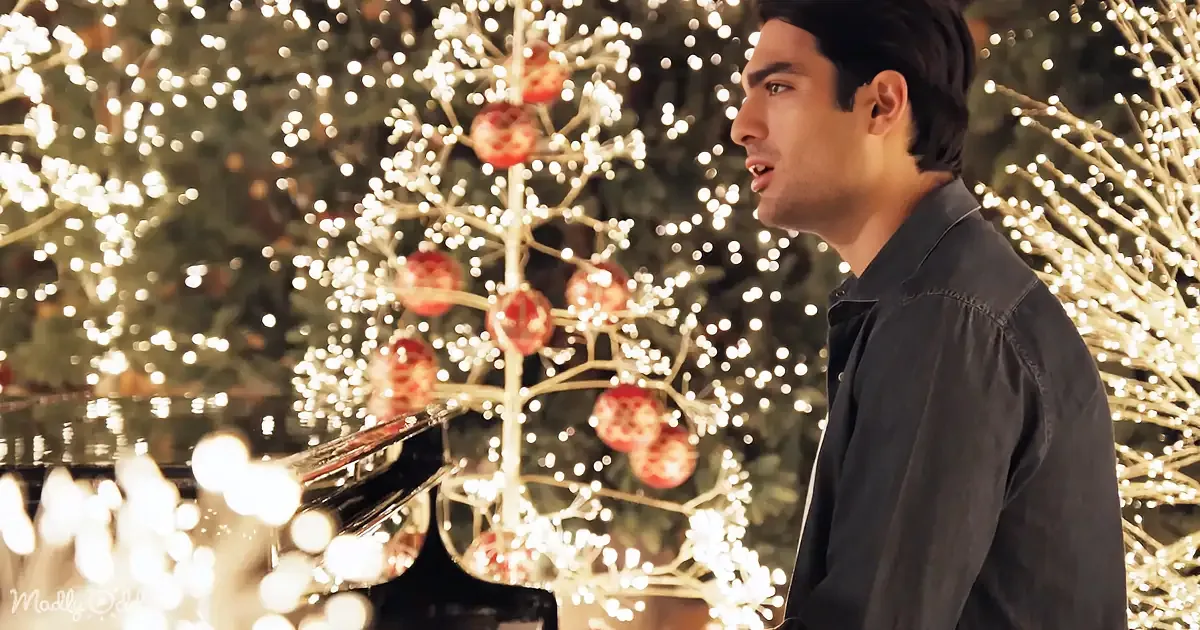 A man in a black shirt stands beside a piano, surrounded by Christmas lights and ornaments.