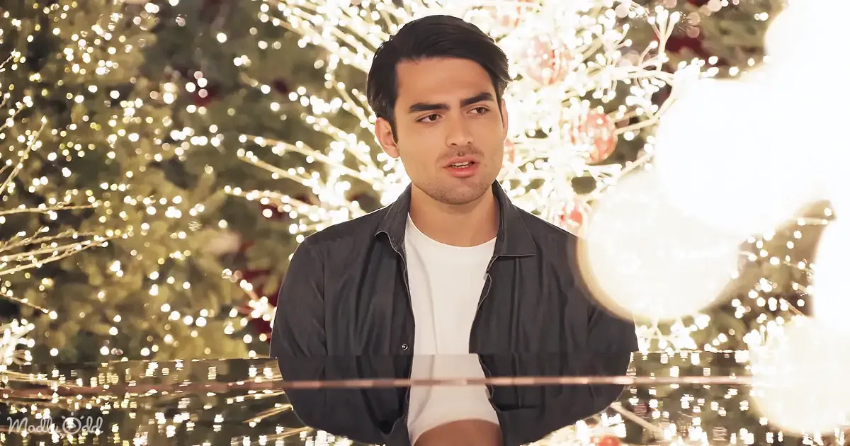 A man in a dark jacket and white shirt stands in front of a brightly lit Christmas tree.