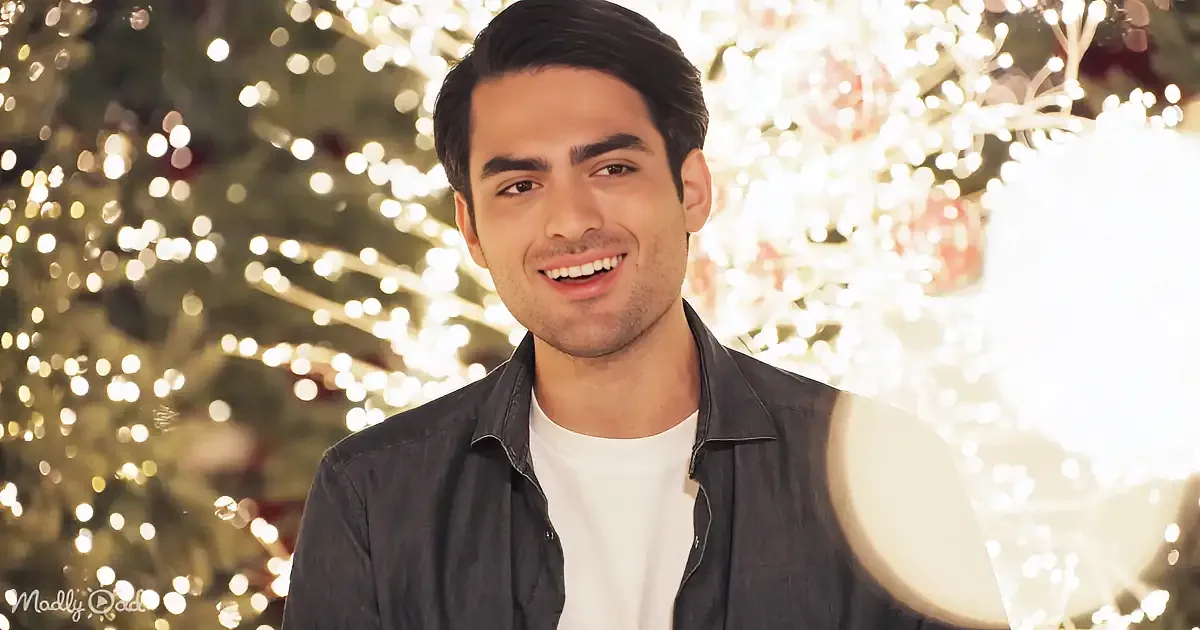 A smiling man in a denim shirt stands in front of a Christmas tree adorned with lights.