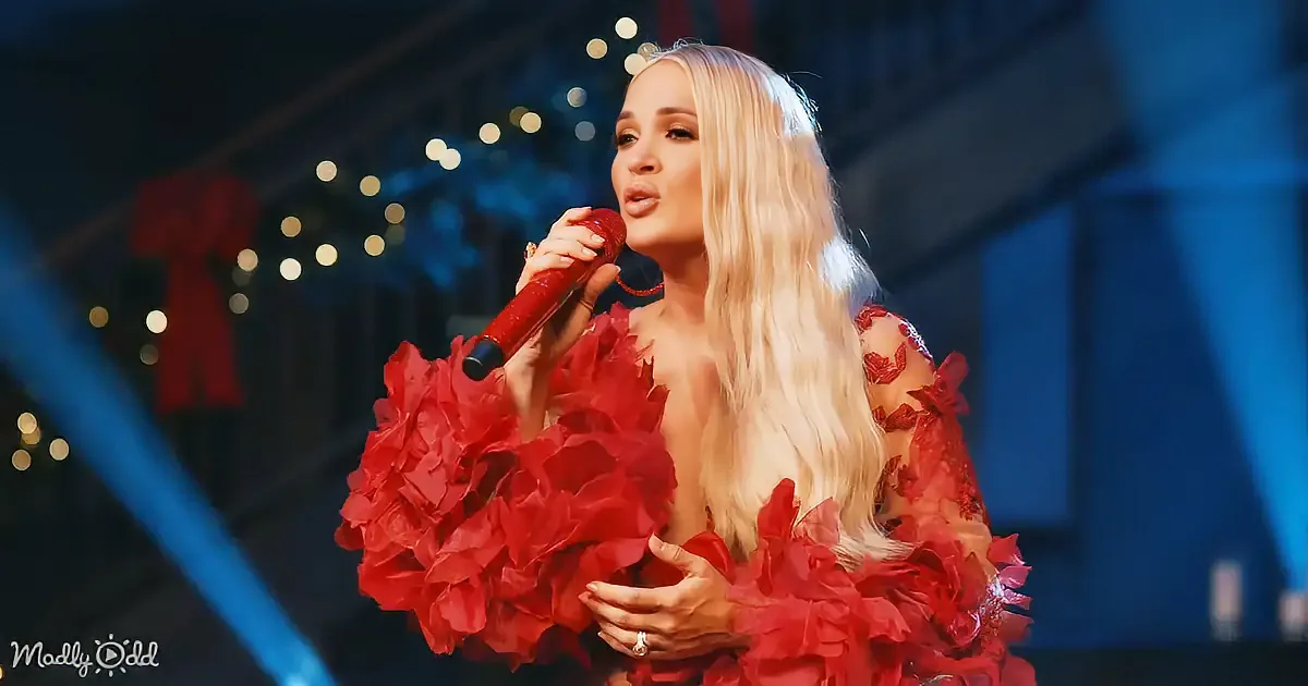 A woman with long blonde hair in a red floral dress sings into a microphone on stage.