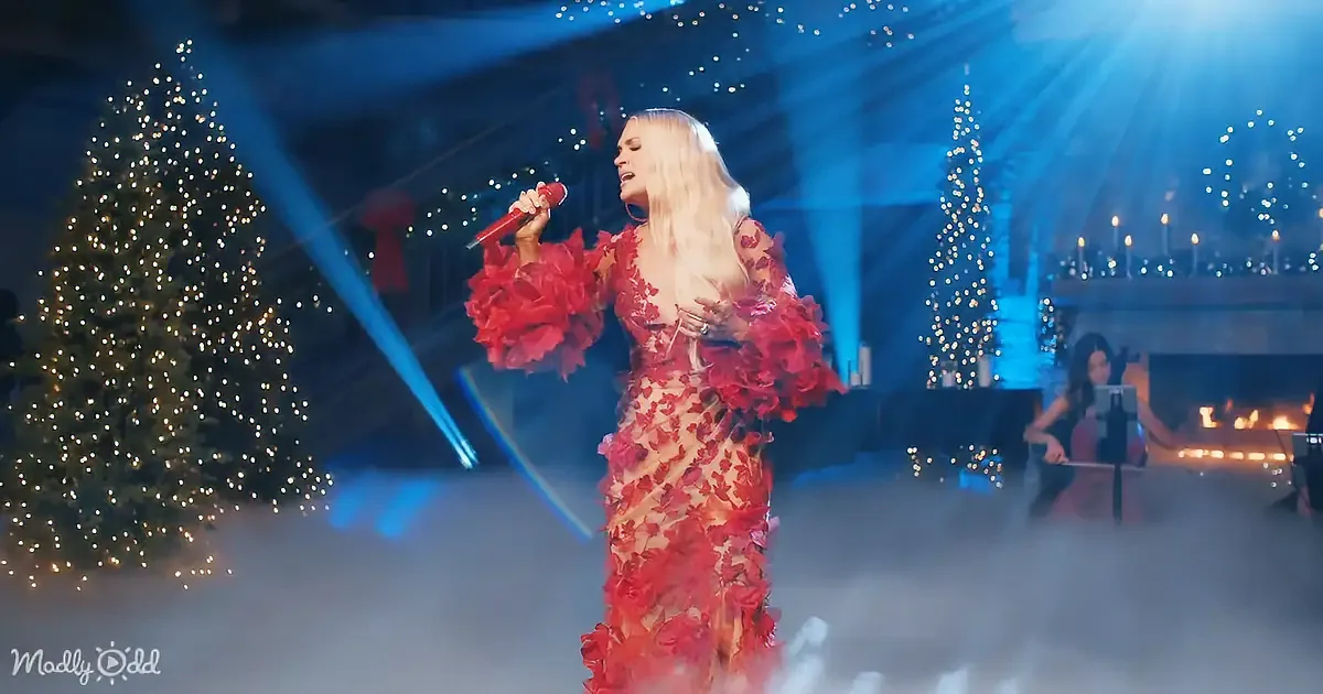 A woman in a red floral dress sings on stage with Christmas trees and blue lights in the background.