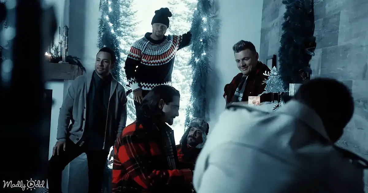 A man in a red plaid shirt and black scarf is seated, surrounded by others in winter attire, in a snowy, festive setting.