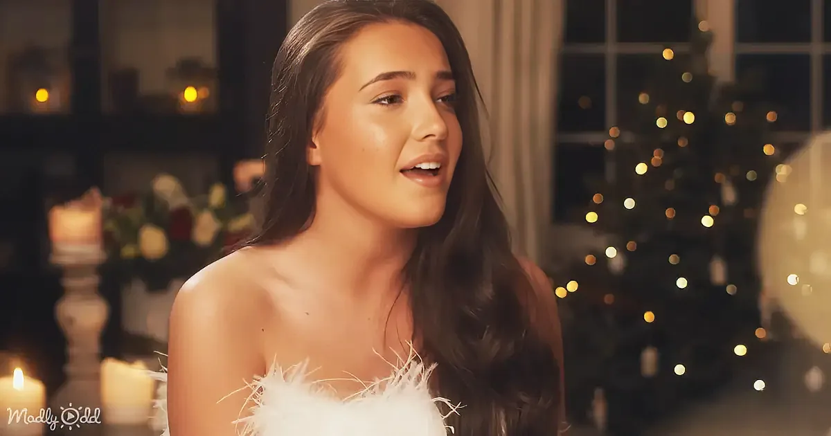 A woman with long dark hair, wearing a white feathered top, sings in a warmly lit room with a Christmas tree and candles.