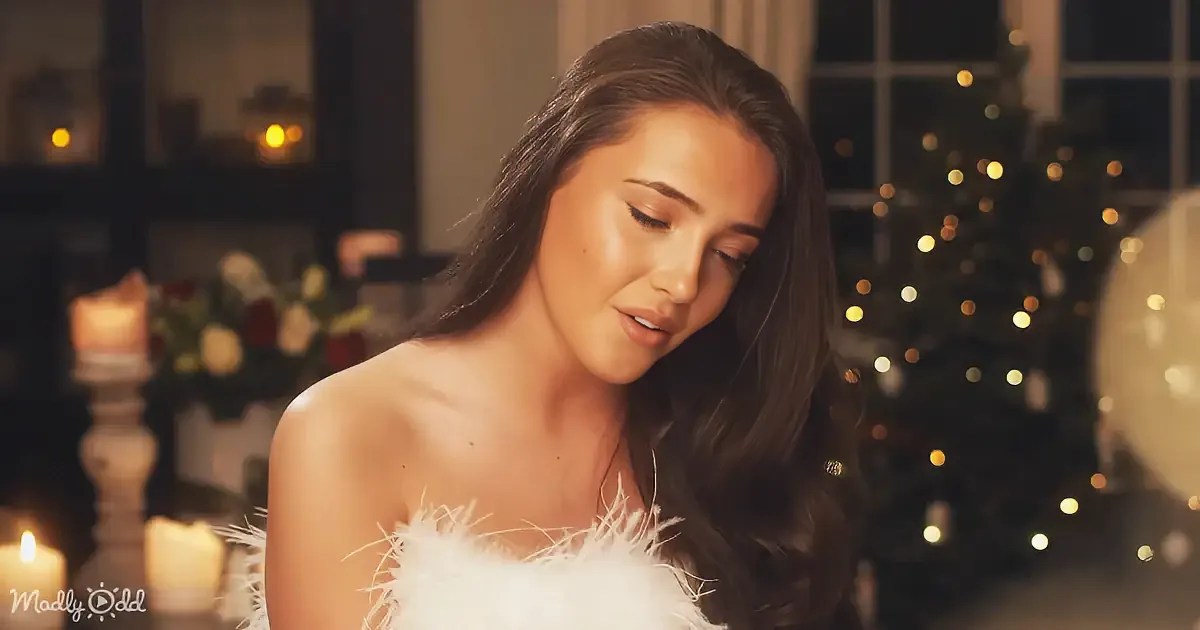 A woman with long dark hair, wearing a white feathered top, is looking down in a warmly lit room with a Christmas tree...