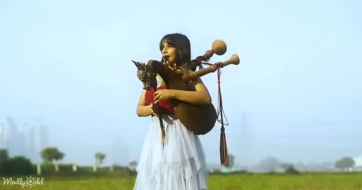 A person in a white skirt plays a bagpipe-like instrument in a grassy field with a cityscape in the background.