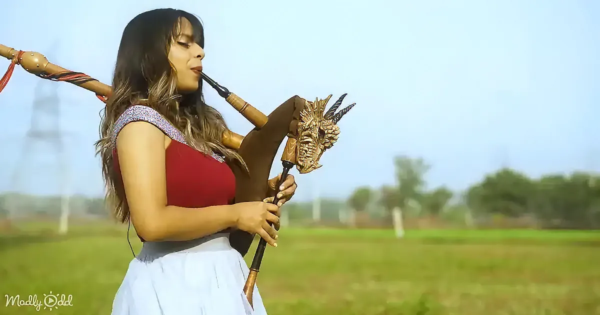 A woman in a red top and white skirt plays a bagpipe with a dragon head, standing in a grassy field.