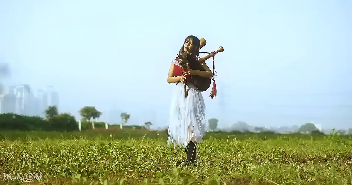A woman in a red top and white skirt plays a bagpipe in a grassy field with a cityscape in the background.