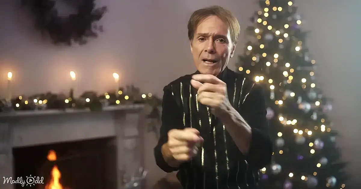 A man in a black sequined shirt stands in front of a Christmas tree and fireplace, gesturing with his hands.