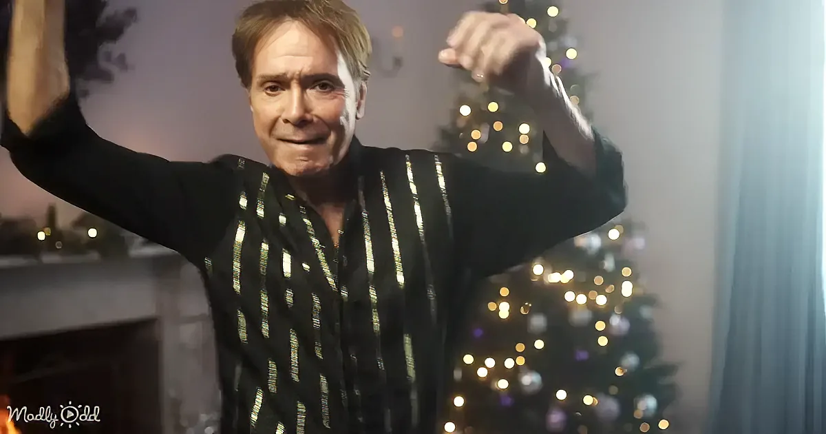A man in a striped shirt raises his arms, smiling, with a Christmas tree and lights in the background.
