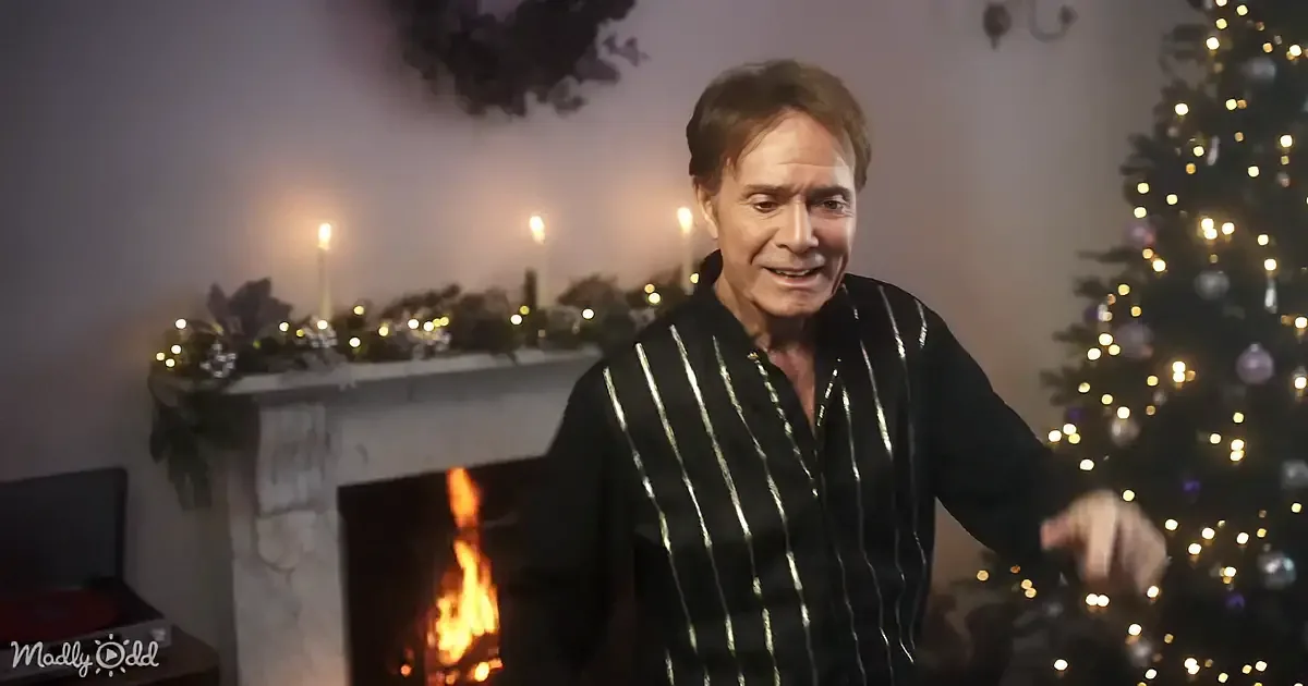 A man in a striped shirt stands near a fireplace and Christmas tree, smiling.