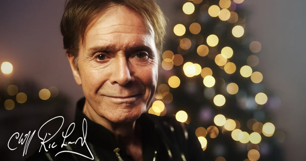 A smiling man in a black jacket with blurred Christmas lights in the background.