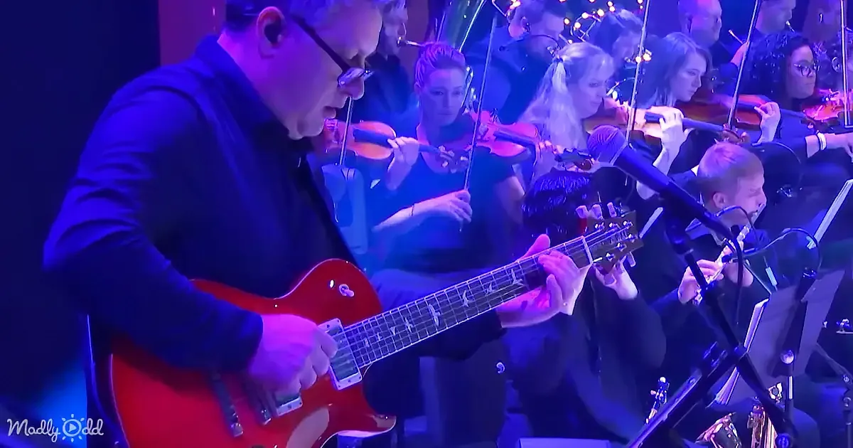 A man in a black shirt plays a red electric guitar on stage with a microphone and orchestra in the background.