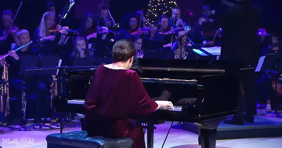 A woman in a maroon dress plays the piano in front of an orchestra under purple lighting.