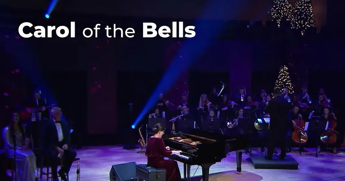 A woman in a red dress plays a grand piano on stage, with a microphone and audience in the background.