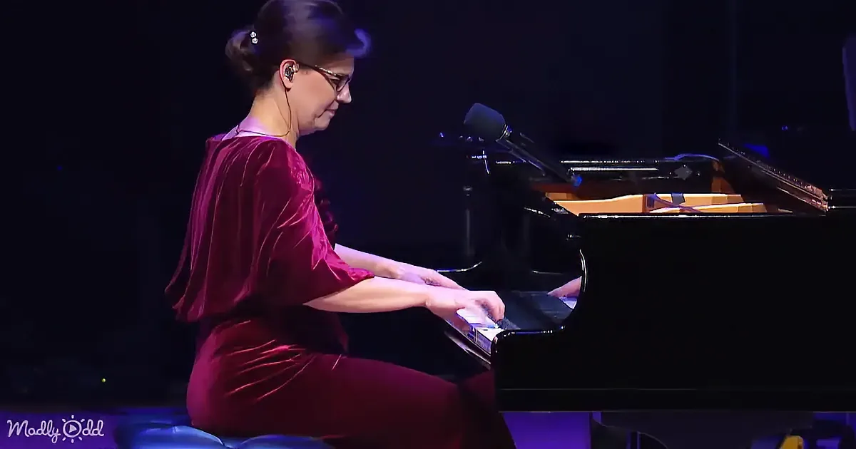 A woman in a red dress plays a grand piano on stage under purple lighting.