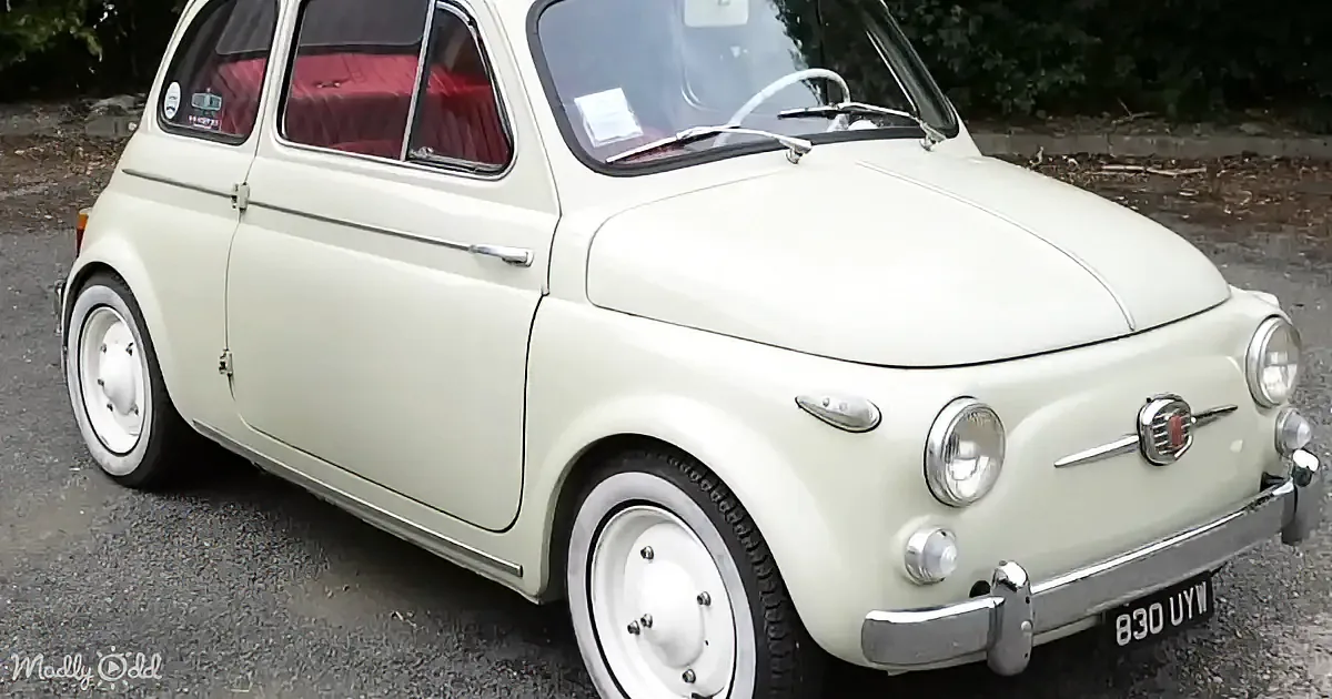 A vintage cream Fiat 500 with white wheels and red interior parked on gravel.