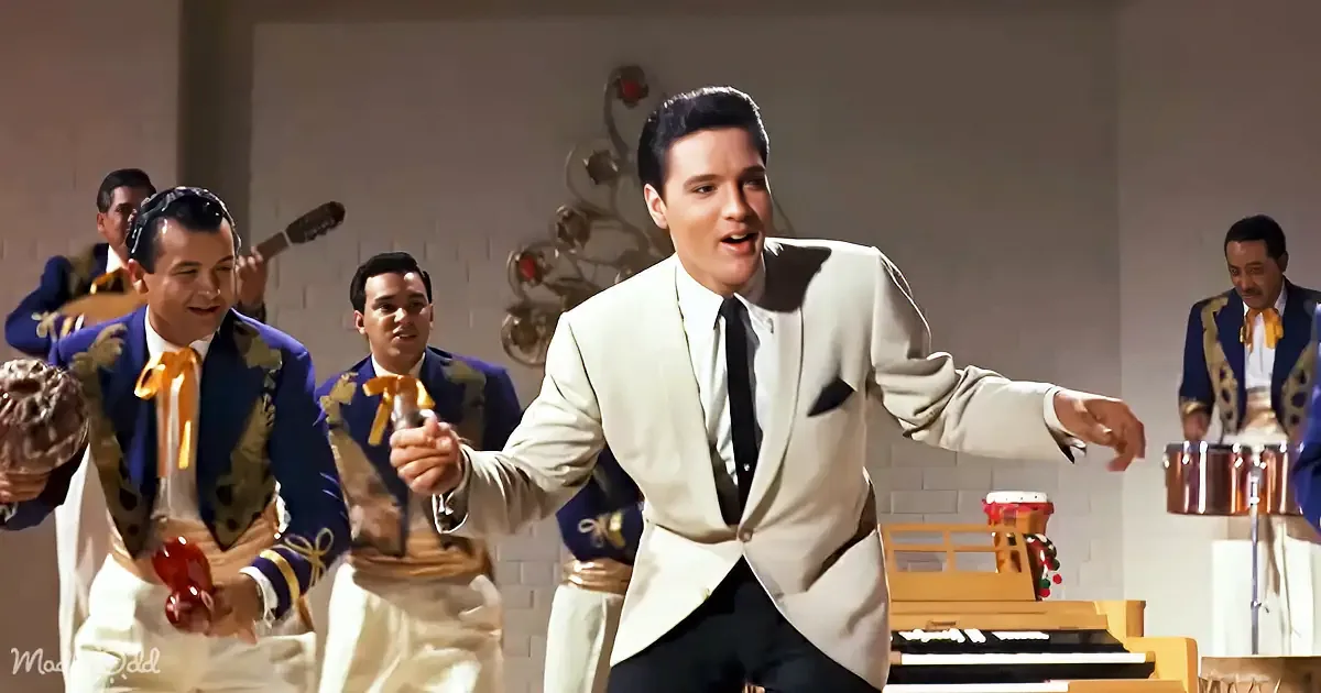 Elvis Presley in a white suit dances energetically, surrounded by musicians in colorful costumes.