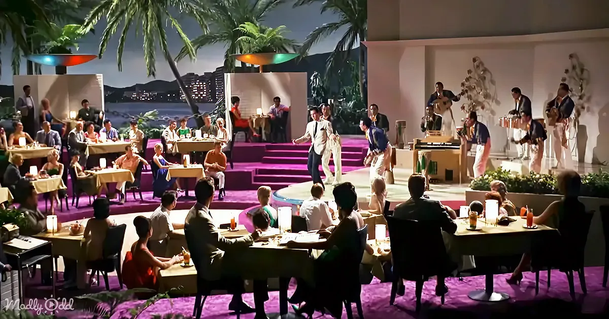 A man in a white suit dances on a stage in a vibrant, tropical-themed restaurant with patrons enjoying the performance.