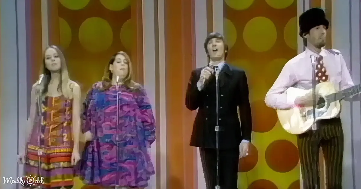 A man in a suit sings on stage with two women in colorful dresses and a guitarist in a hat.