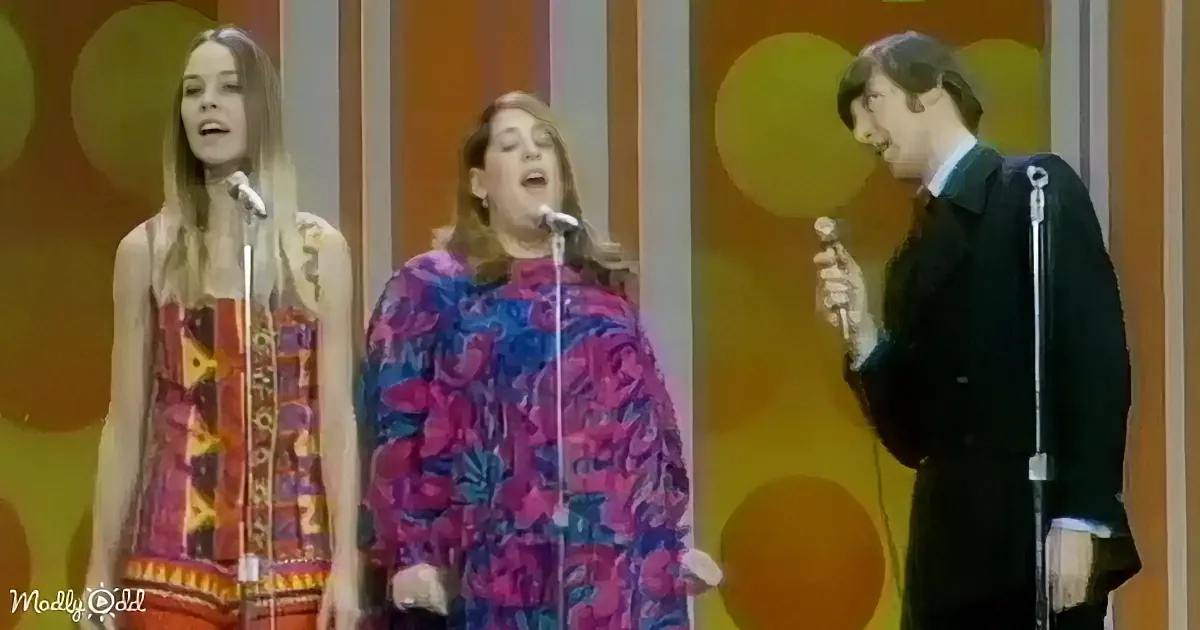 Three performers sing on stage, two women in colorful outfits and a man in a suit, against a vibrant, retro backdrop.