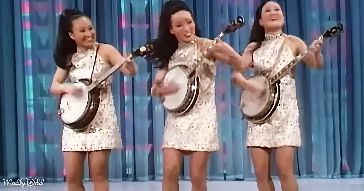 Three women in white dresses play banjos on a stage with a blue curtain backdrop.