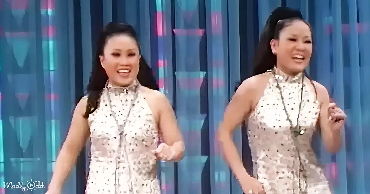Two women in matching sequined dresses are dancing on a stage with a colorful backdrop.