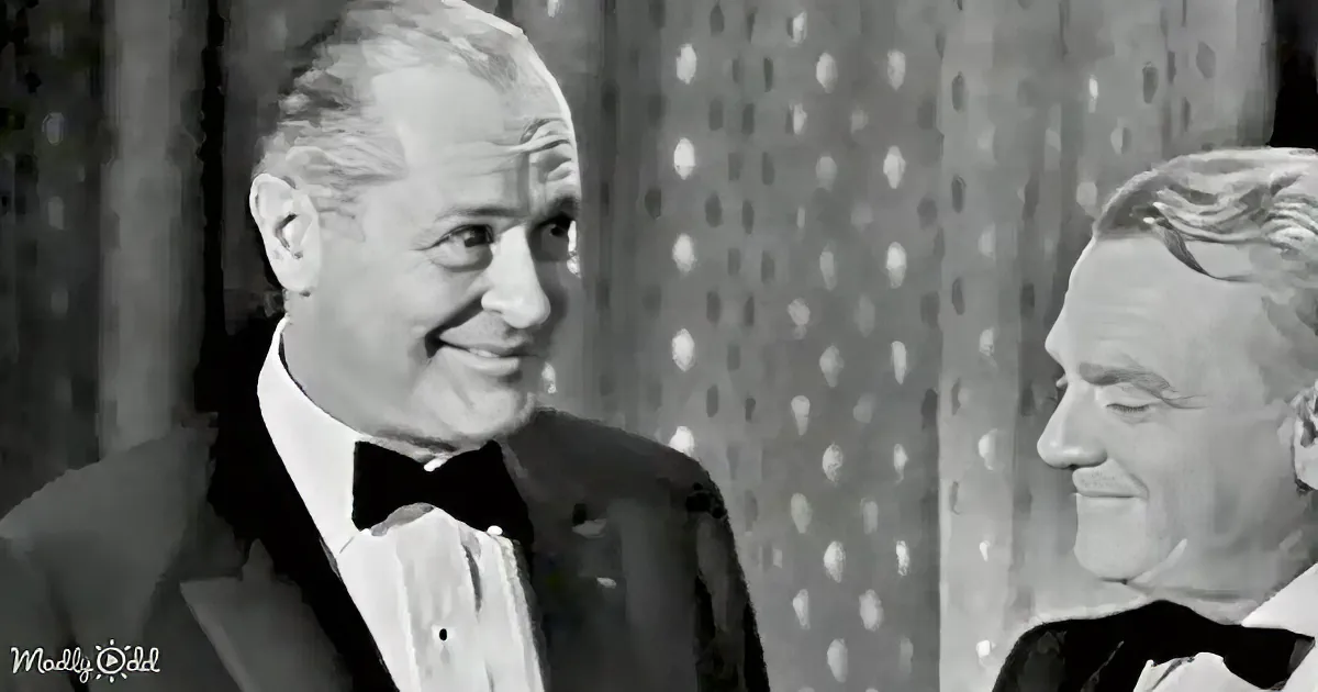 A man in a tuxedo smiles warmly at another man in a tuxedo, both against a patterned backdrop.