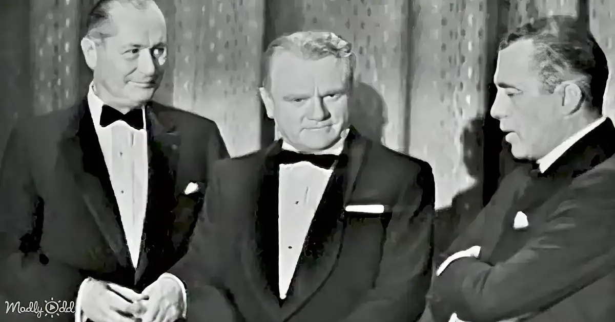 Three men in tuxedos stand together, one in the center appears to be speaking or reacting to something.