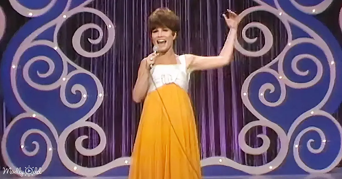 A woman in a white top and yellow skirt sings on stage with a microphone, surrounded by swirling blue and white backdrop.
