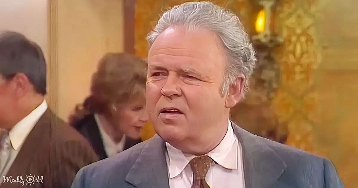 A man in a suit and tie looks to the side with a serious expression, surrounded by other people in a formal setting.