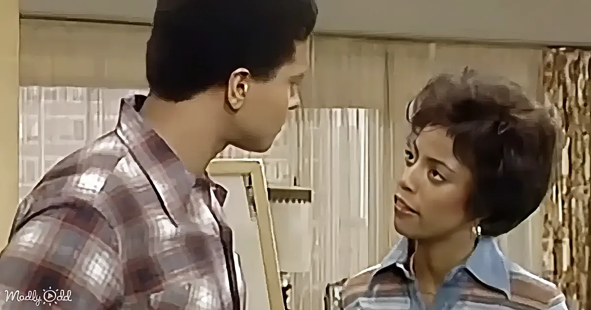 A man in a plaid shirt and a woman in a striped shirt are facing each other in a room with curtains.