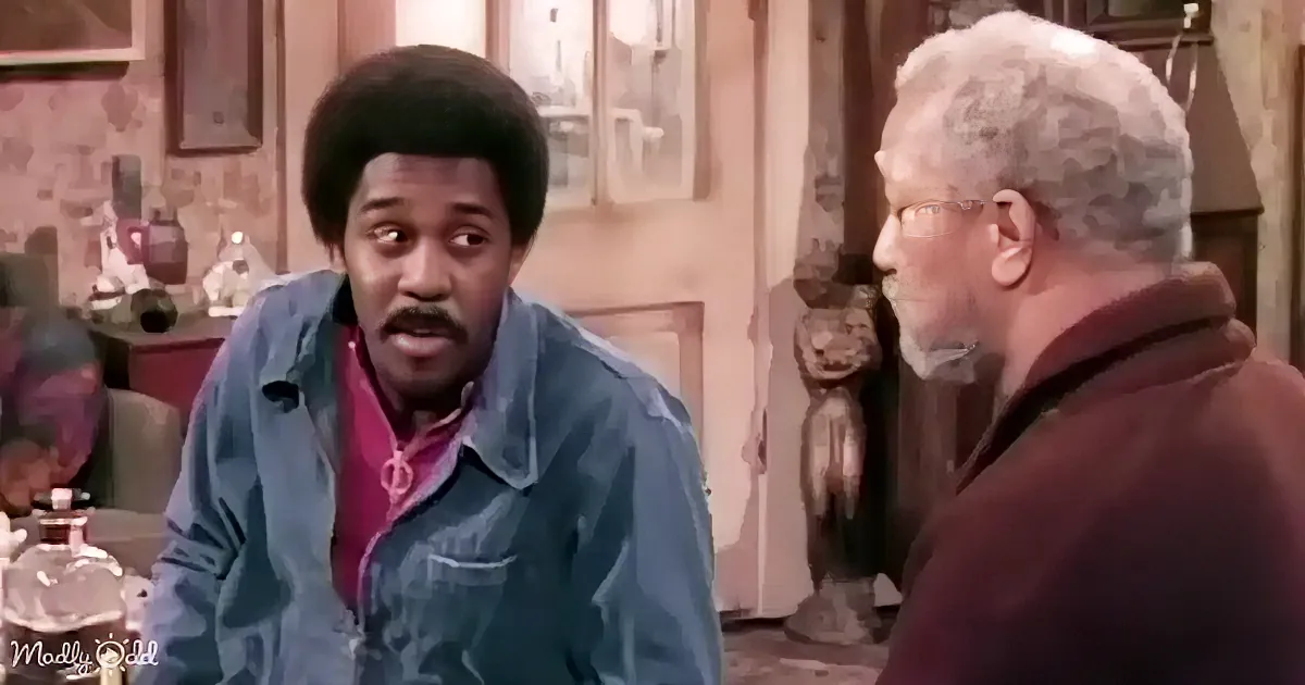 A man in a blue jacket is speaking to another man in a brown jacket in a room with artwork and a bottle on a table.