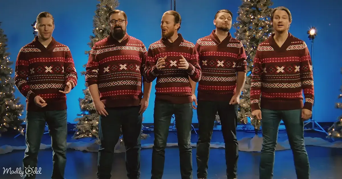 Five men in matching red Christmas sweaters sing in front of decorated trees and a blue backdrop.
