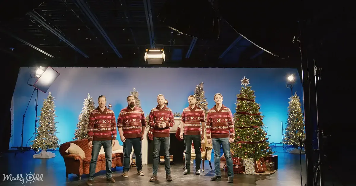 Five men in matching sweaters stand in front of a Christmas-themed backdrop with trees and lights.
