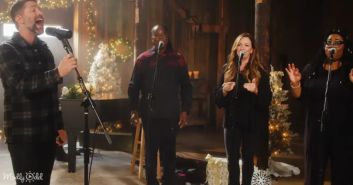 A group of four people singing in front of Christmas decorations, with a man in a plaid shirt leading.