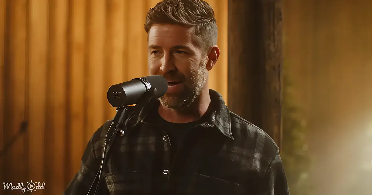 A man with a beard speaks into a microphone, wearing a plaid shirt, against a wooden backdrop.