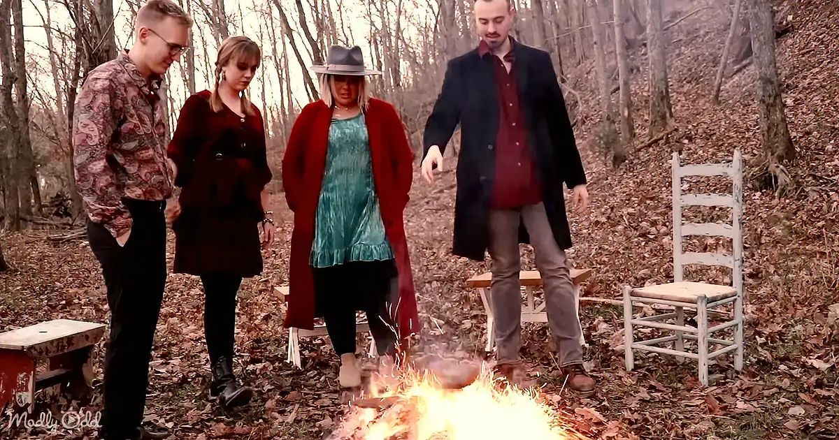 Four people stand around a fire in a forest, dressed in coats and casual attire.