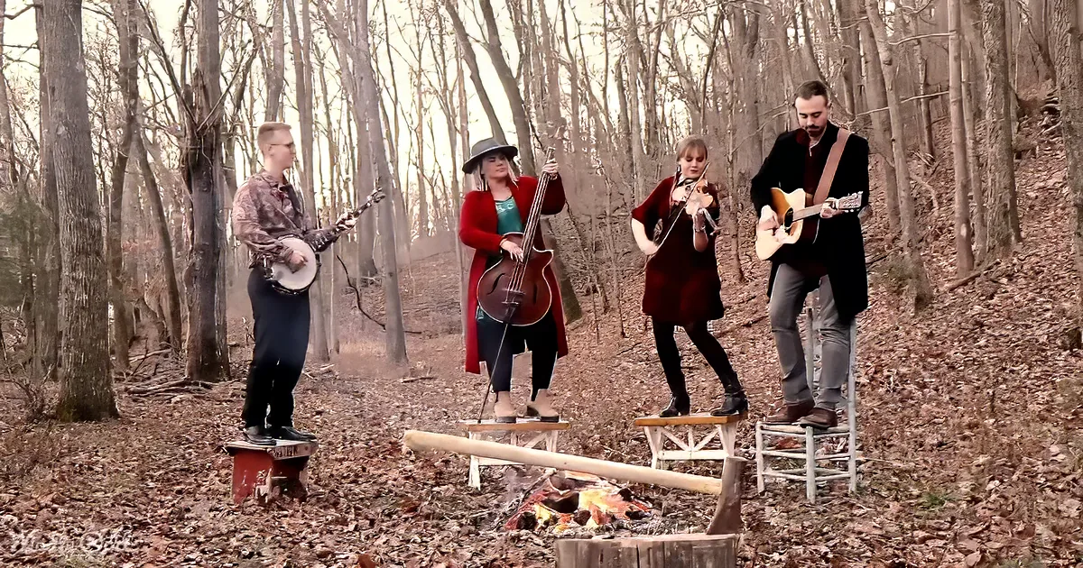 A group of musicians plays instruments in a forest setting near a campfire.