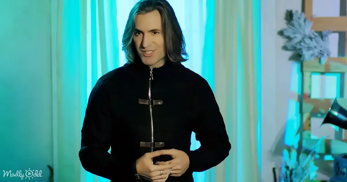 A person with long hair, wearing a black jacket with a zipper, stands in front of a blue-lit background.