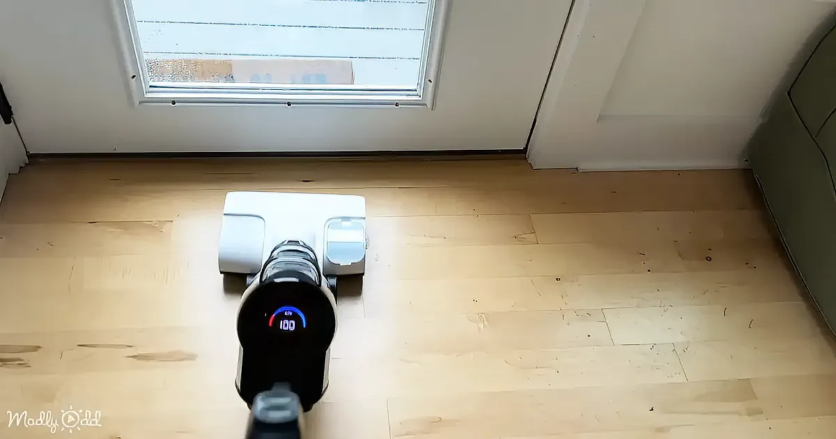 A robot vacuum cleaner is on a light wooden floor near a window and a door.