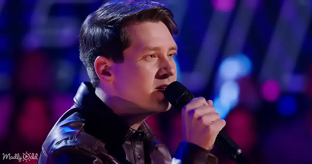 A man in a leather jacket sings into a microphone on a stage with colorful lights.