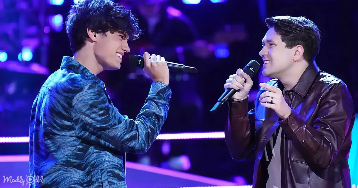 Two men in a stage setting, one in a blue jacket holding a microphone, the other in a brown jacket, both smiling and...