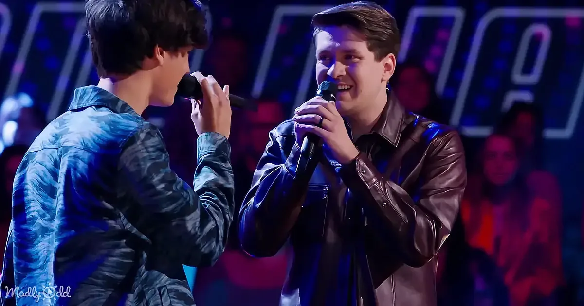 Two young men sing on stage, one in a blue jacket, the other in a brown leather jacket, holding microphones.