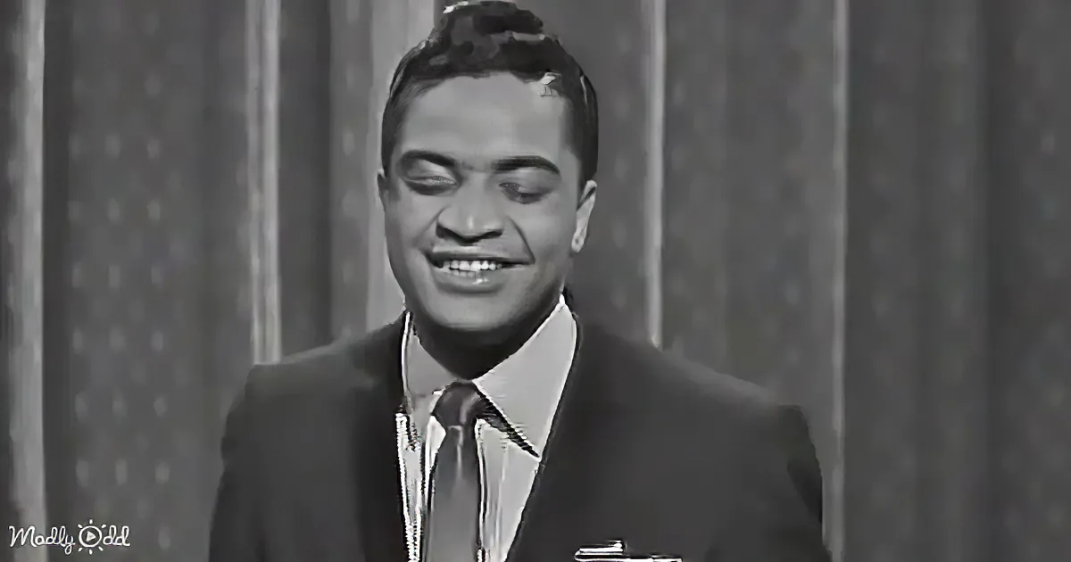 A man in a suit and tie smiles, eyes closed, against a patterned backdrop.