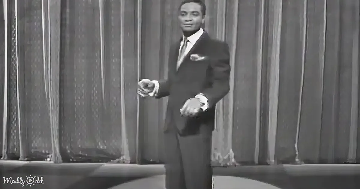 A man in a suit and tie stands on a stage, hands clasped, against a curtain backdrop.