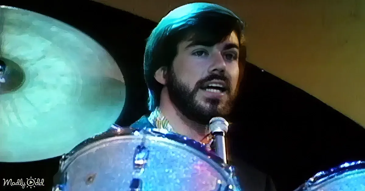 A man with a beard and dark hair is playing drums, singing into a microphone.