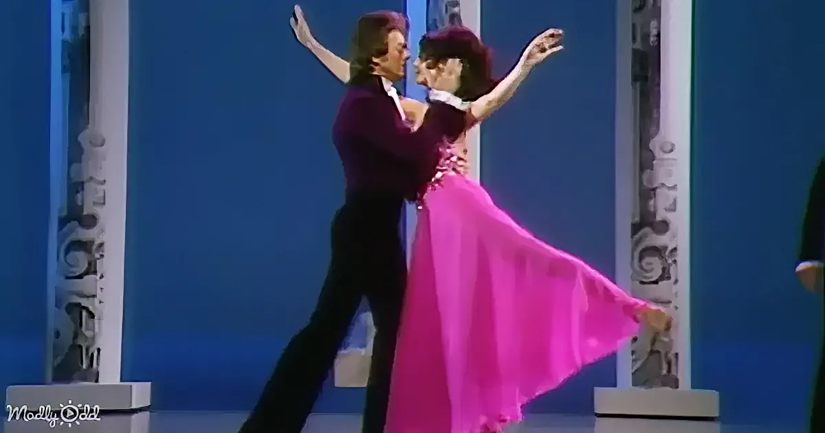 A couple in formal attire dances gracefully on a stage with a blue backdrop.